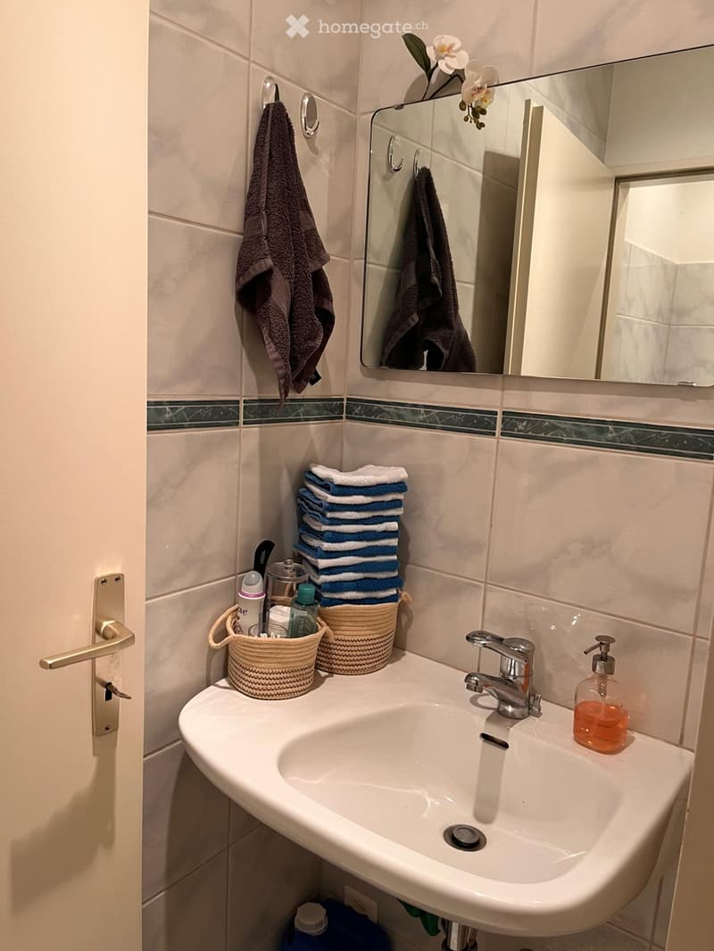 A modern bathroom with a white sink, chrome faucet, mirror, and neatly stacked towels. A soap dispenser and baskets with toiletries are placed on the sink counter.