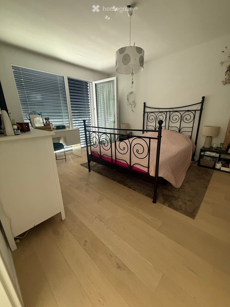 Bright bedroom with wooden flooring, large windows with blinds, a metal-framed bed with pink bedding, a nightstand, a small table, and a hanging lamp.