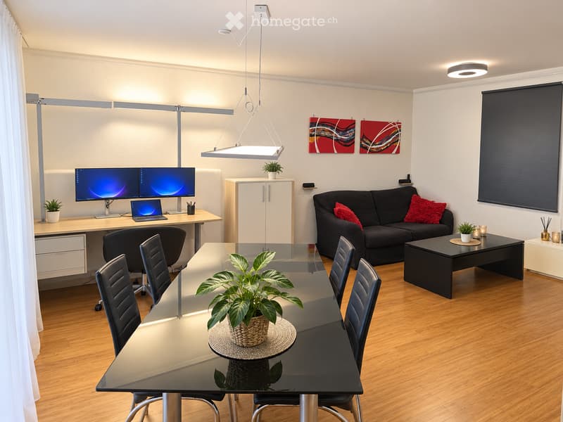 Modern living and dining area with a black sofa, red cushions, coffee table, dining table with chairs, laptop, and two screens on a desk.
