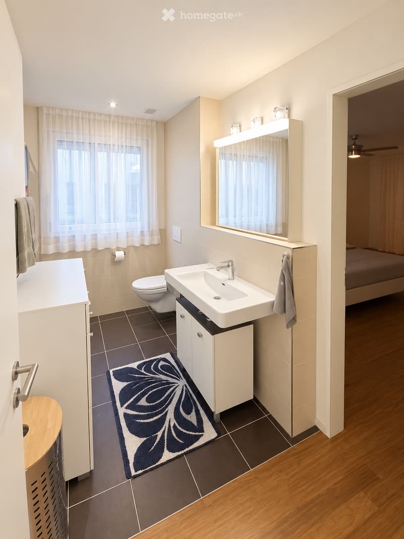 Modern bathroom with a white sink, toilet, bathtub, large mirror, and a decorative rug. The space features light-colored tiles and a wooden floor, with a towel hanging on the wall and a small cabinet beneath the sink.