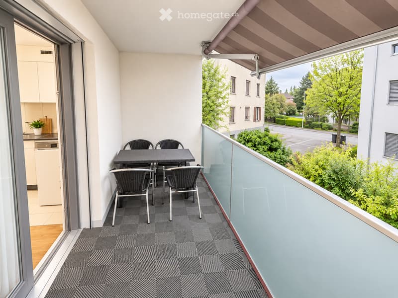 Balcony with a black dining table and four chairs, glass railing, and a view of greenery and neighboring buildings.