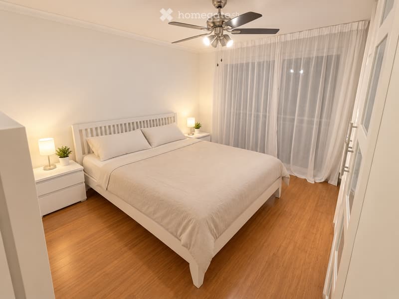 Cozy bedroom with a white bed, nightstands, lamps, and a ceiling fan. The room features wooden flooring and large windows with white curtains.