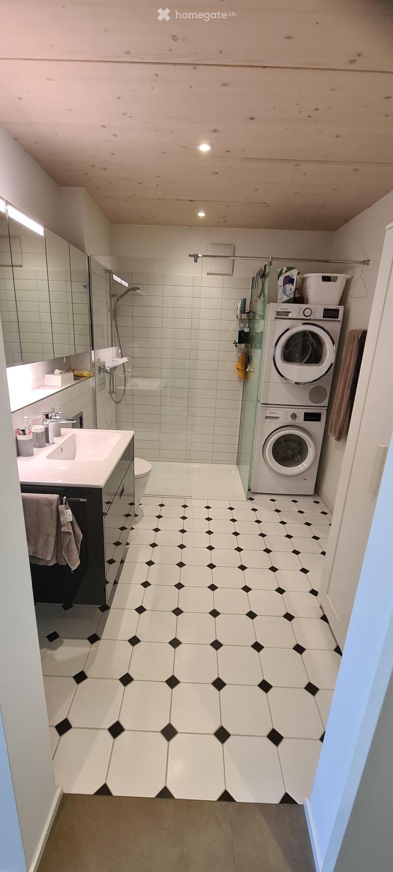 Modern bathroom with white tiles, hexagonal floor tiles, a sink, toilet, shower, and a stacked washing machine and dryer.