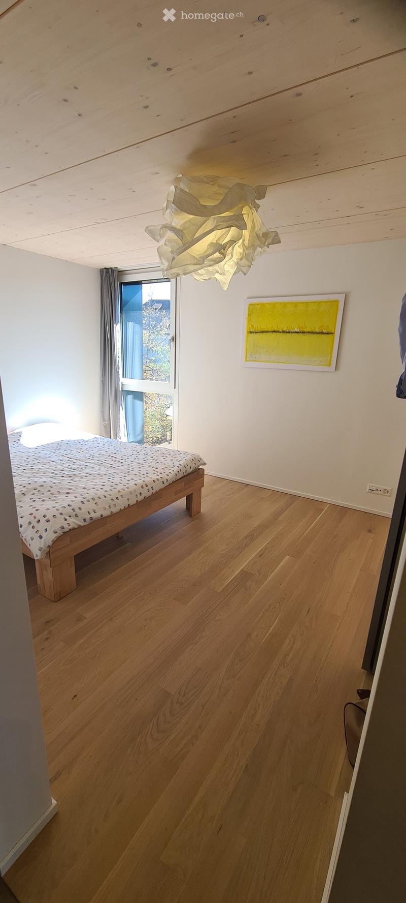 Bright bedroom with wooden flooring, a single bed with patterned bedding, a modern abstract painting, and a unique hanging light fixture.