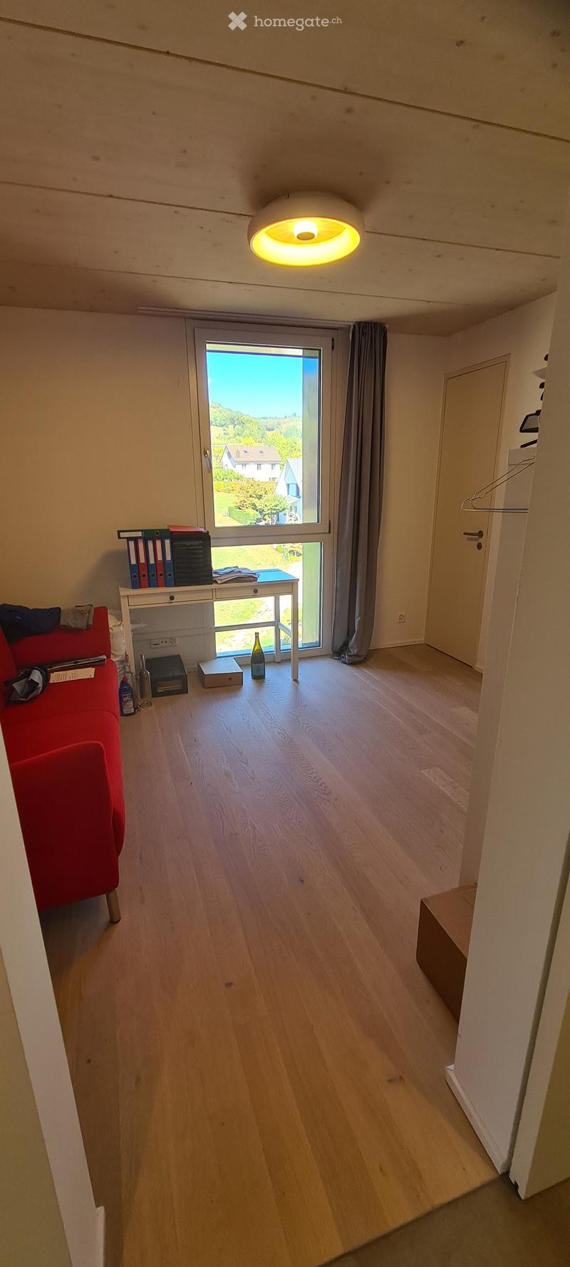 Bright room with wooden flooring, large window with view, red sofa, small table with books and bottles, and a hanging lamp.