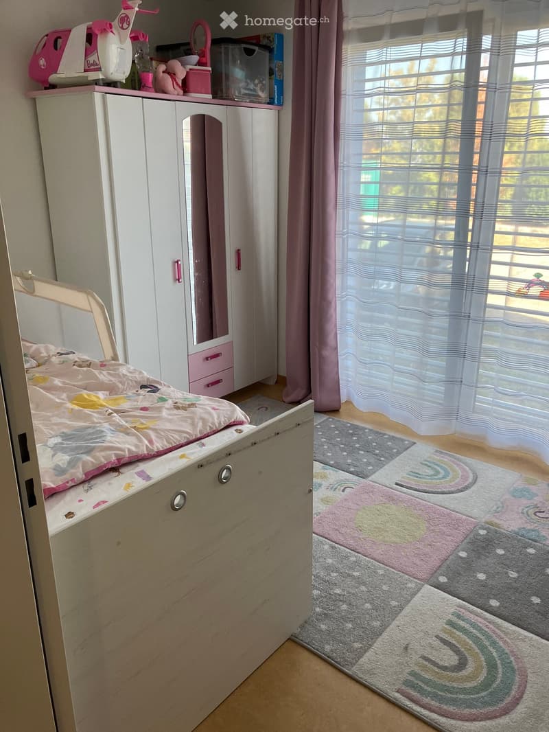 A child’s bedroom with a bed, colorful bedding, a wardrobe with a mirror, a toy on top, and a rainbow-patterned rug.