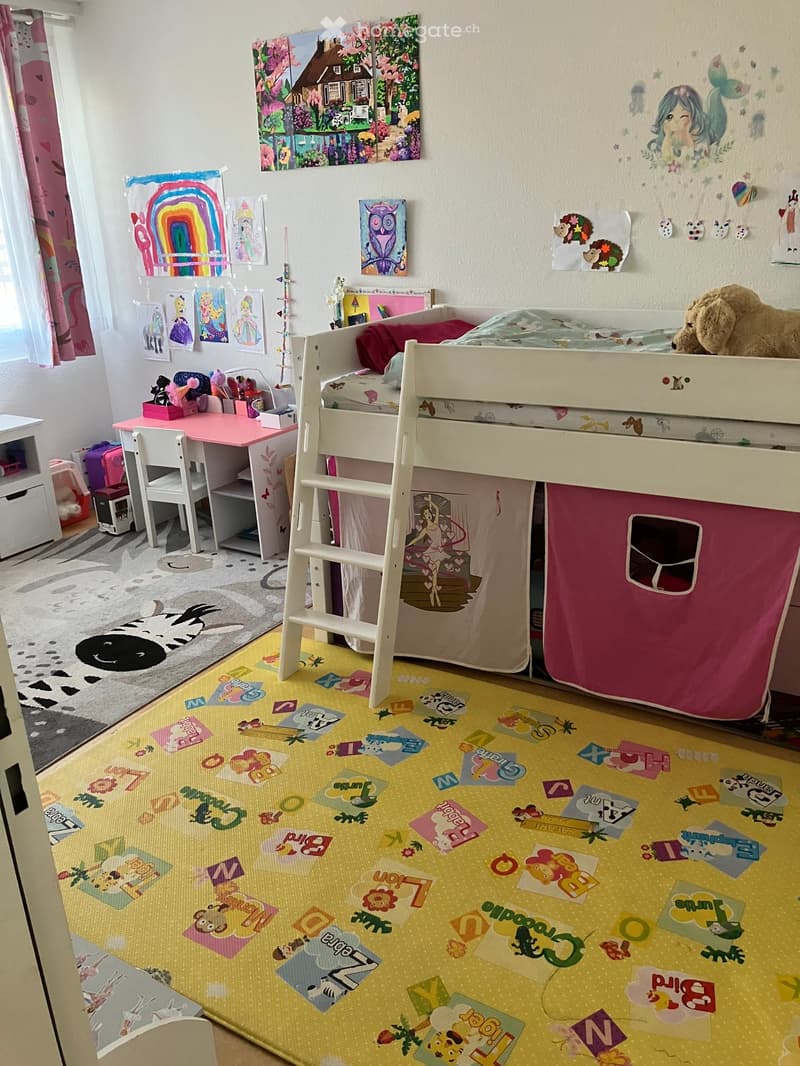 Child's bedroom with a loft bed, desk, chair, and colorful artwork on the walls. The room features a vibrant yellow rug with playful patterns and a cozy stuffed animal on the bed.