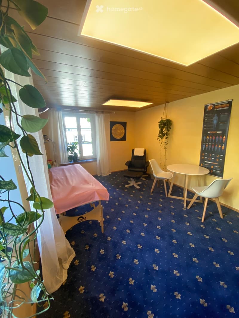 A well-lit room with a massage table, chairs, a small table, and a poster on the wall.