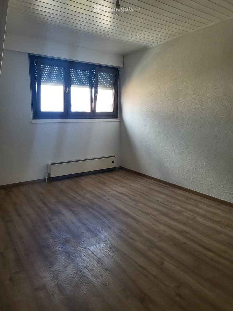 Empty room with wooden flooring, white walls, and a large window with blinds. A radiator is visible under the window.