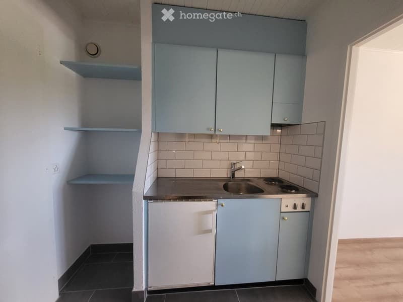 Compact kitchen with light blue cabinets, stainless steel sink, and electric stove. White tiled backsplash and dark tiled floor.