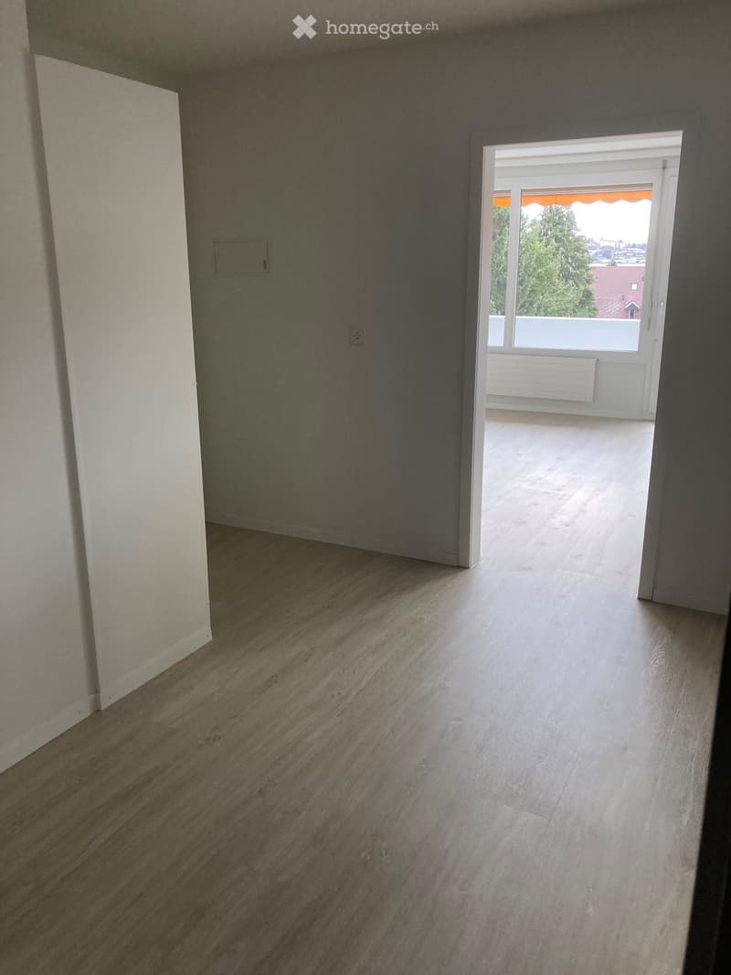 Empty room with light wooden flooring and white walls, featuring an open doorway leading to a balcony with a view of trees and a building.