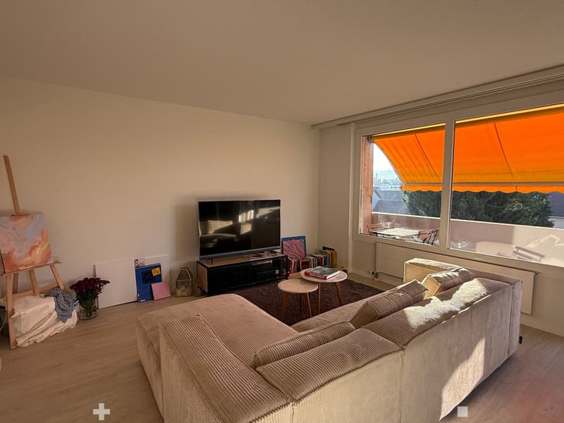 Modern living room with a large window, a grey sofa, a coffee table, a television, a small round table, and decorative items including a vase with flowers and an easel with a canvas.