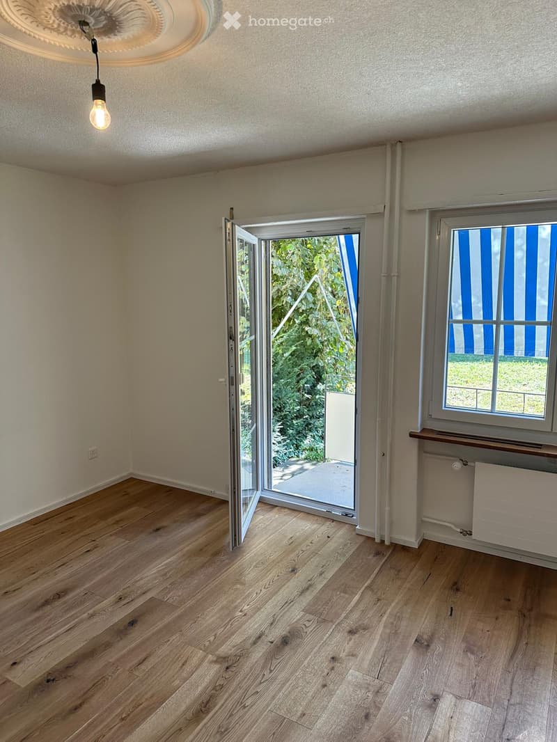 Empty room with wooden flooring, large sliding glass door leading to outdoor area, window with blue blinds, radiator, and light fixture on the ceiling.