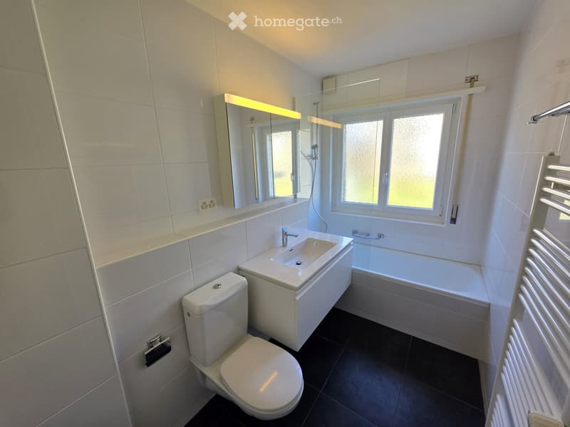 Modern bathroom with white tiles, a toilet, a sink with a mirror, a bathtub, and a radiator. The room has a window with frosted glass and a built-in shelf above the sink.