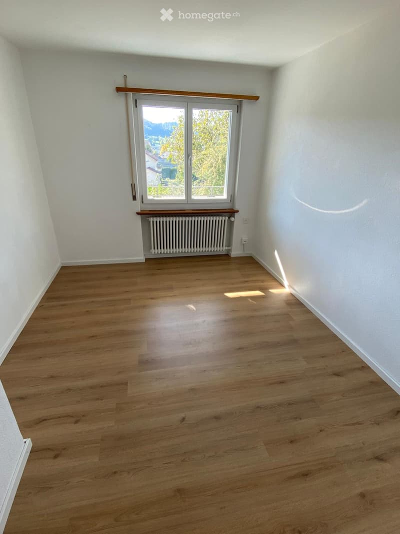 Empty room with wooden flooring, white walls, a radiator, and a large window with a view of greenery.