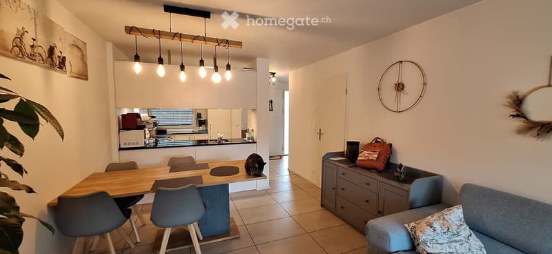 Open-plan living and dining area with a grey sofa, coffee table, dining table with chairs, kitchen counter with appliances, wall clock, and decorative items.