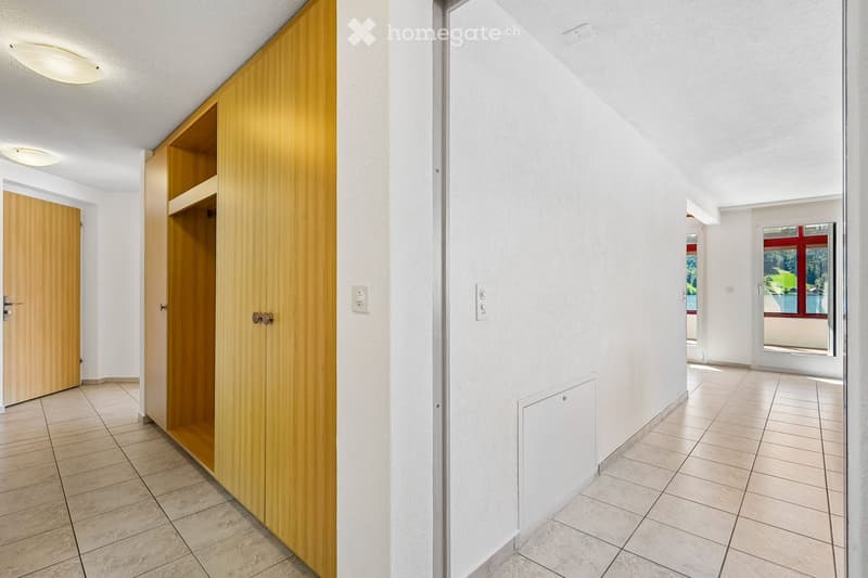 Clean, modern hallway with tiled flooring, white walls, and wooden cabinetry. Features recessed lighting, electrical outlets, and doors leading to other rooms.