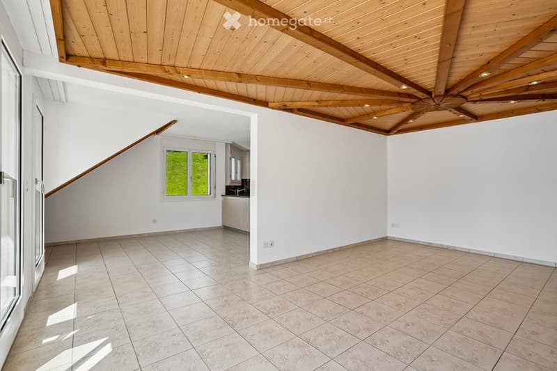 Empty room with white walls, tiled floor, and wooden ceiling beams. Features a staircase leading to another level and a small window with a view of greenery.