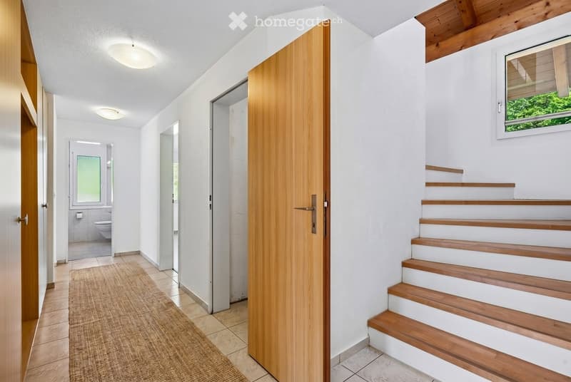Modern hallway with wooden staircase, white walls, and recessed lighting. Features a wooden door, a small rug, and a glimpse of a bathroom through an open doorway.