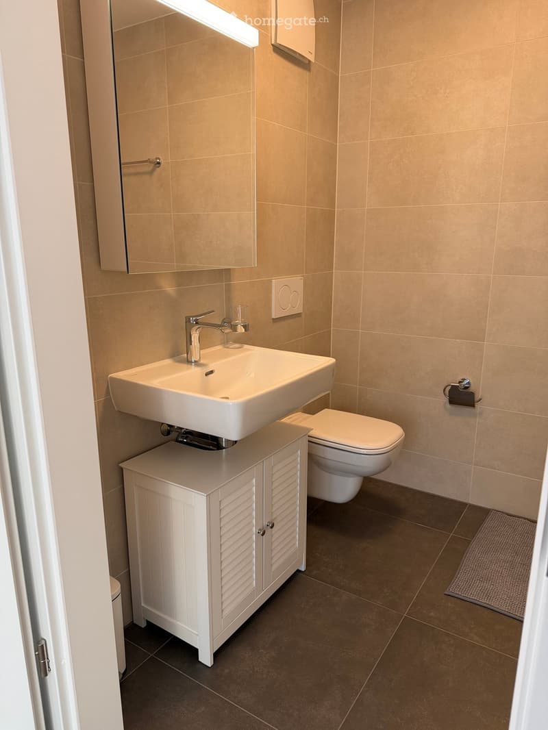 Modern bathroom with beige tiled walls, a white sink with a chrome faucet, a white cabinet, a toilet, and a mirror with built-in lighting.