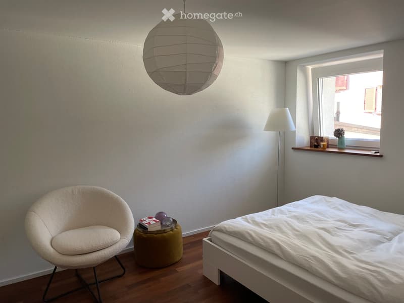Modern bedroom with a white bed, round chair, small table, and a hanging paper lantern light fixture. A window with a small shelf holds decorative items.
