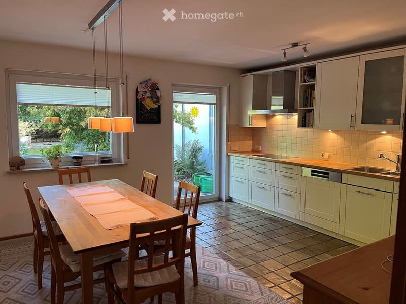 Bright kitchen with white cabinets, a wooden dining table with chairs, and a sink area with a faucet and dishwasher. Natural light streams in from large windows and a sliding door leading to a garden.