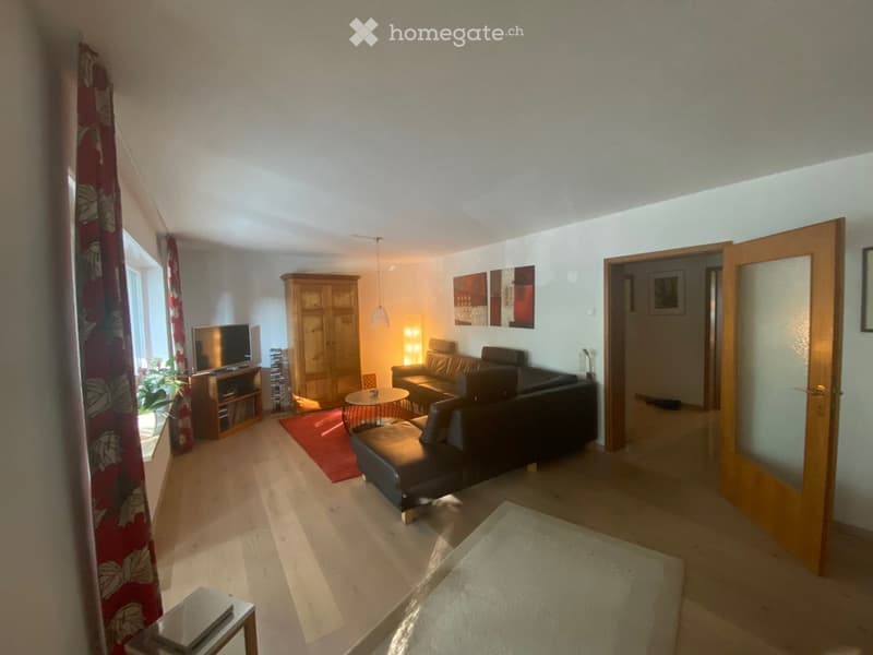 Modern living room with a leather sofa, coffee table, TV, wooden cabinet, and a small round table with a chair. The room features large windows with curtains, a red rug, and a white shag rug on the floor.