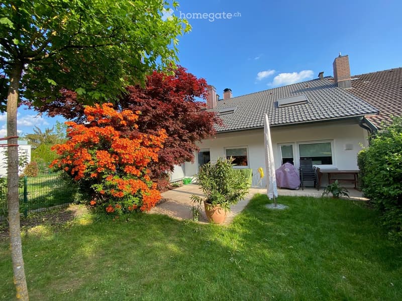 A well-maintained garden with a lawn, colorful flowering shrubs, a patio area with outdoor furniture, and a modern house in the background.