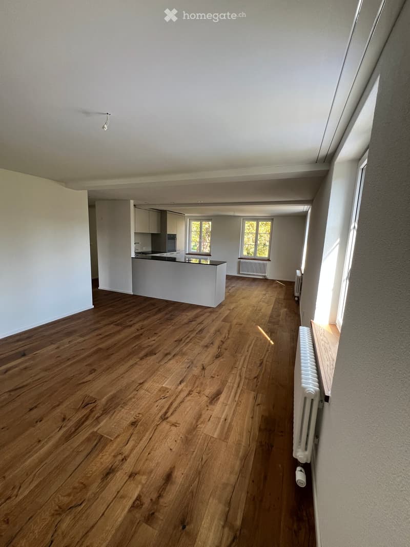 Empty room with wooden flooring, white walls, and a small kitchen area visible in the background. No furniture or equipment present.