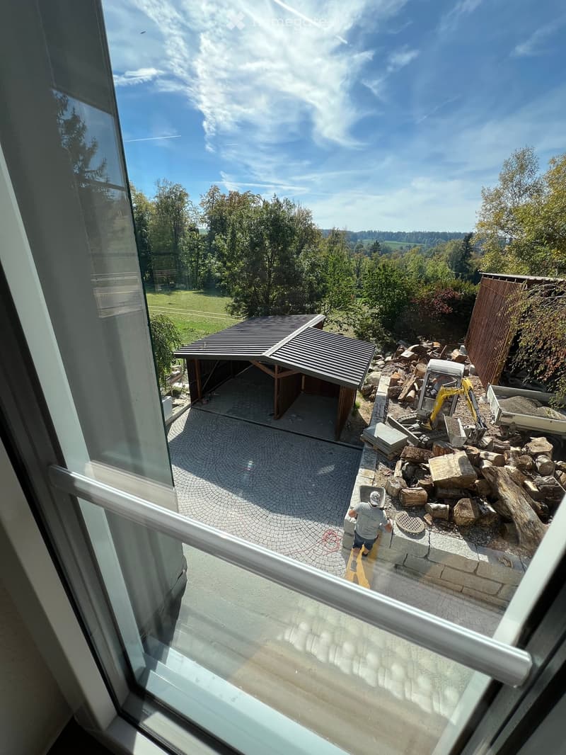 View from a balcony showing a paved area with a wooden shed, construction materials, and a person working with a yellow excavator in the background.