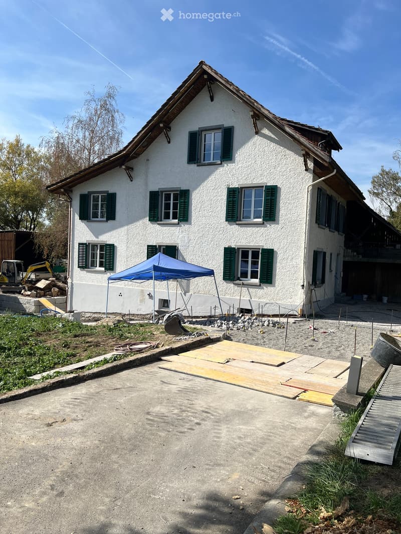 Two-story white house with green shutters and multiple windows, under construction with a blue canopy and construction materials nearby.