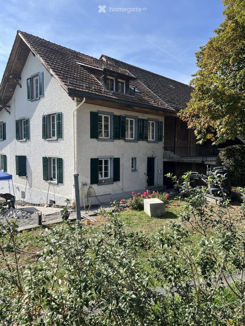 Two-story house with white walls, green shutters, and a tiled roof. Features include a balcony, garage, and garden area with plants and a small construction area.