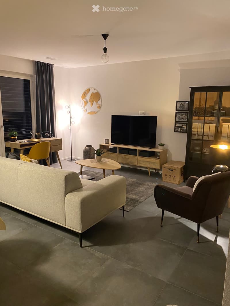 Modern living room with a grey sofa, armchair, coffee table, TV, floor lamp, and a small desk area with a chair and potted plant. Features a world map on the wall and a large window with curtains.