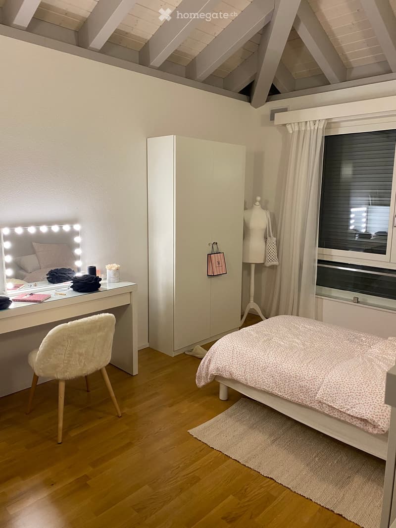 Cozy attic bedroom with wooden slanted ceilings, a bed with a floral duvet, a vanity with a mirror and lighting, a white wardrobe, and a plush chair.