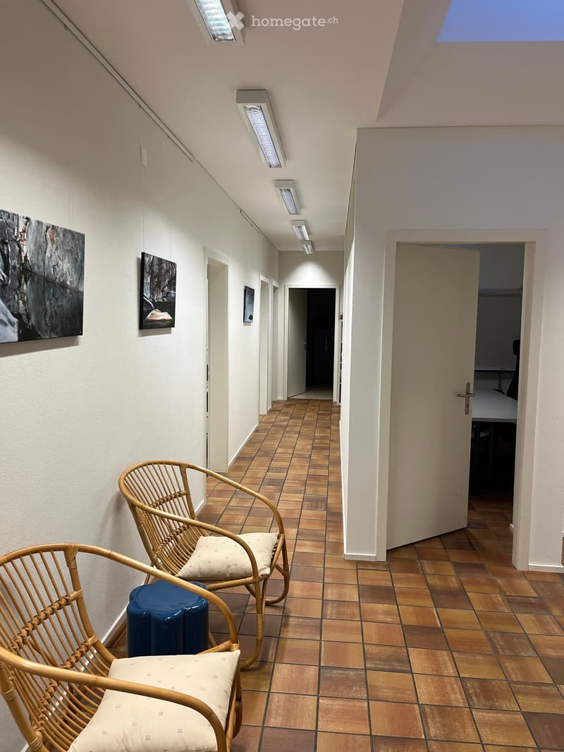 A hallway with tiled flooring, two wicker chairs with white cushions, a blue stool, and several picture frames on the walls.