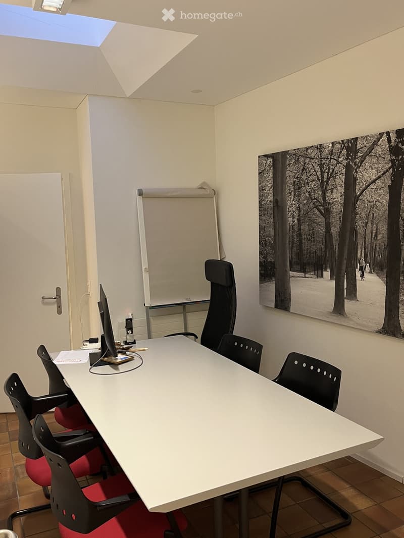 A modern office or meeting room with a large white table, several black chairs with red seats, a screen, a telephone, and a large black-and-white photograph on the wall.