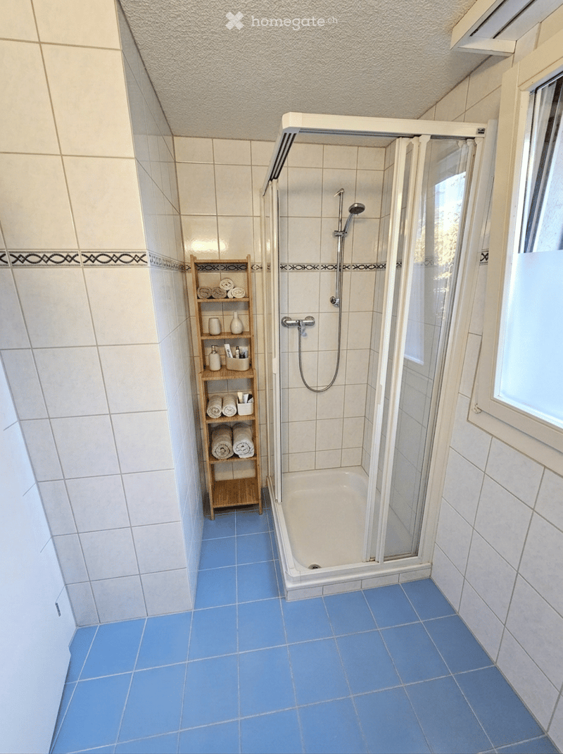 Bathroom with blue tiled floor, white tiled walls, bathtub with glass shower enclosure, wooden shelf with towels and decorative items.