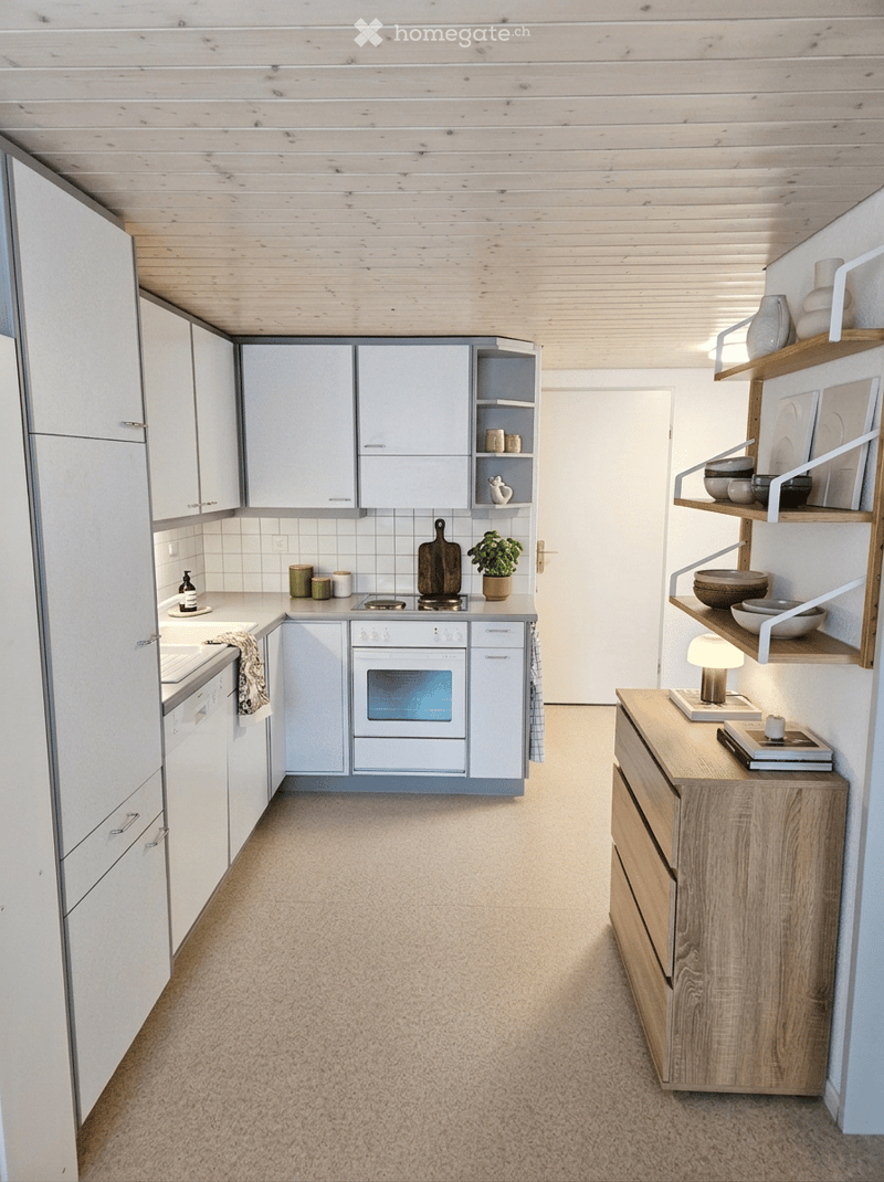Modern kitchen with white cabinets, a built-in oven, a countertop with a cutting board and a small plant, and open shelving displaying bowls and cups. A wooden side table holds a lamp and books.