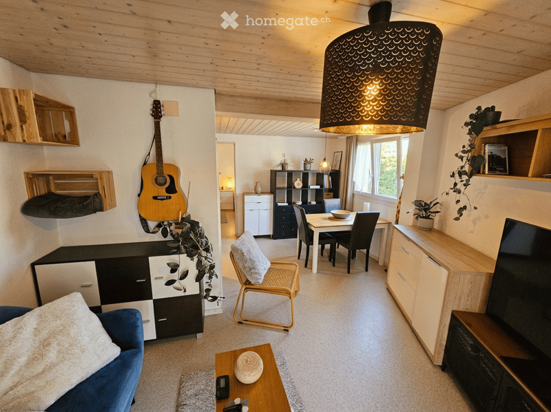 A modern, open-concept living and dining area with wooden ceilings, white walls, and a mix of contemporary and rustic furniture. Features include a blue sofa, a wicker chair, a wooden coffee table, a dining table with chairs, a guitar on the wall, and bui