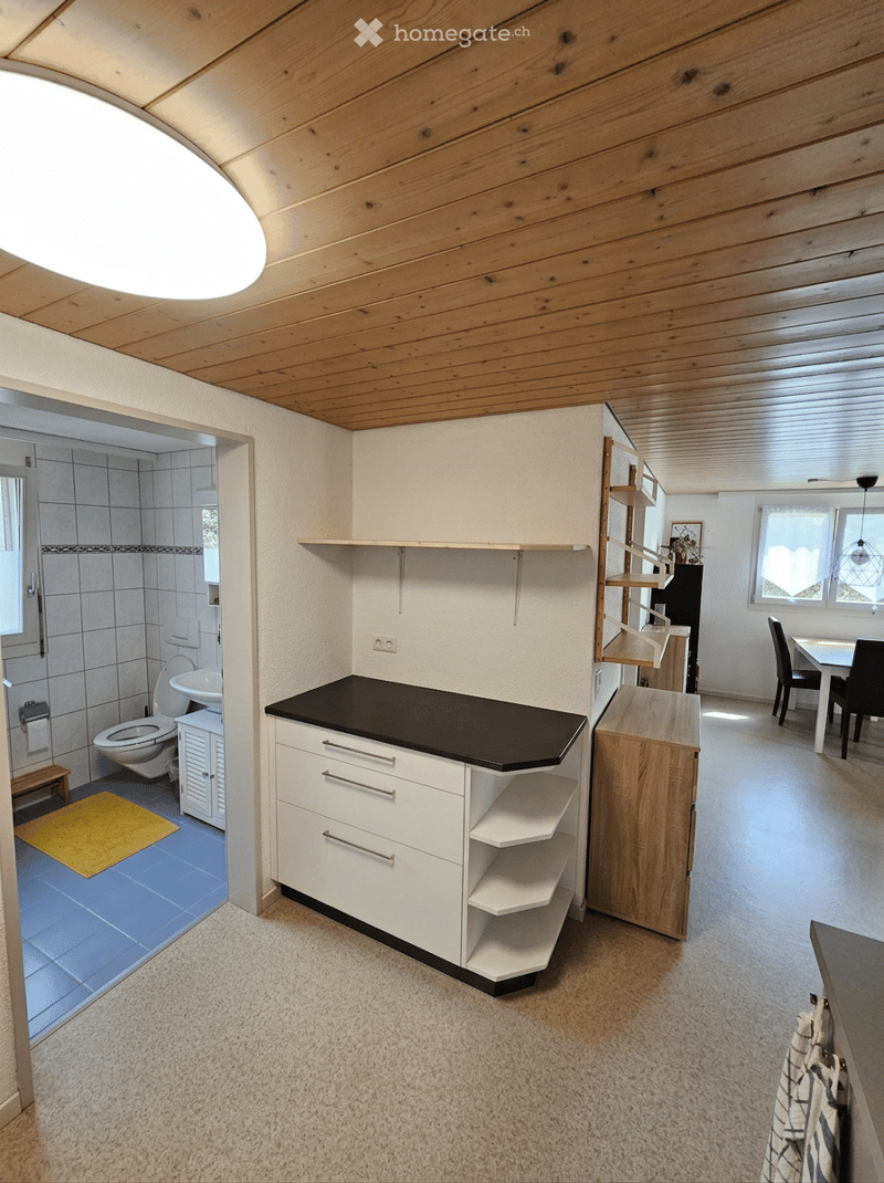 Bright, open-plan room with a kitchen area featuring white cabinets and a black countertop, a dining area with a table and chairs, and a bathroom with a toilet, sink, and mirror. The space has wooden beams on the ceiling and a yellow rug in the bathroom a