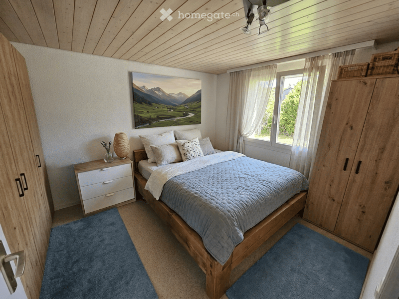 Cozy bedroom with a wooden bed, nightstand, and large window with curtains. Features a scenic mountain view painting on the wall and a wooden wardrobe.