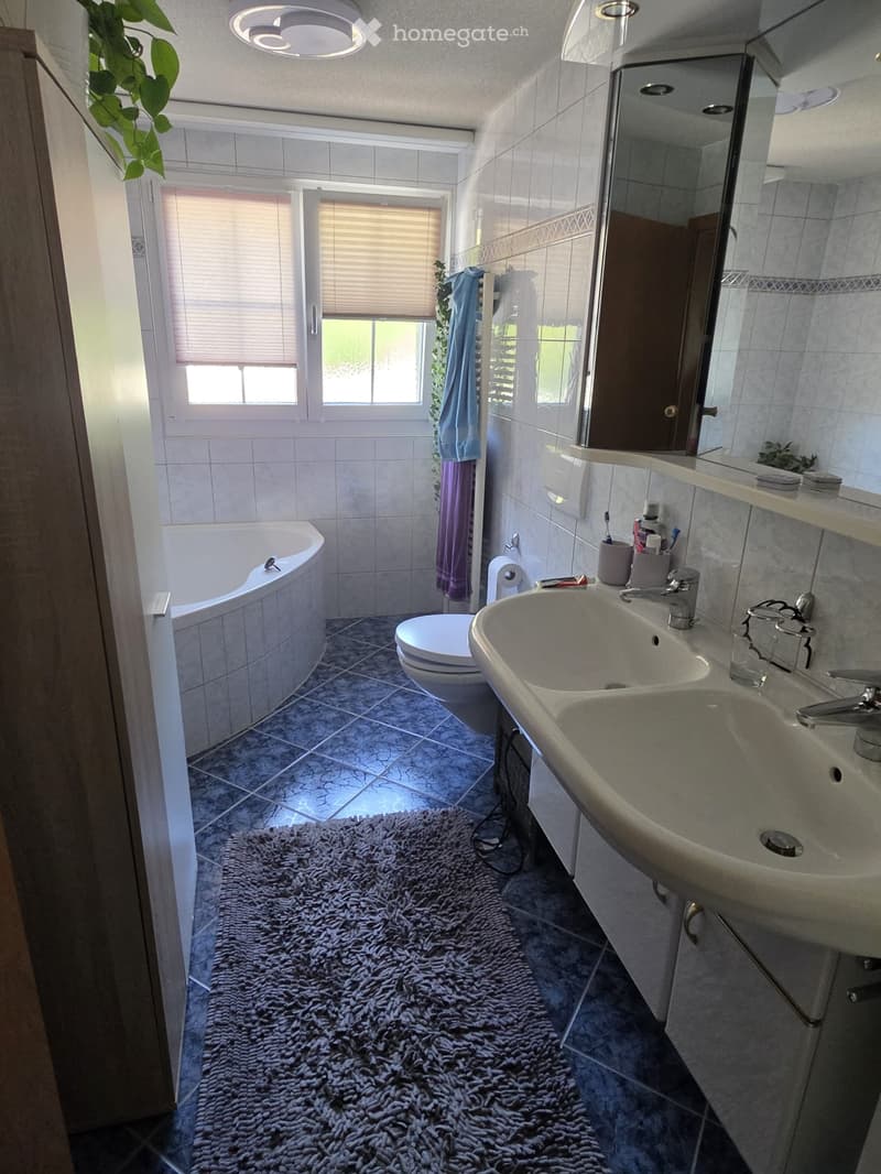 Bathroom with a bathtub, toilet, double sink, mirror, and a fluffy rug on the floor. The room has tiled walls and a window with blinds.