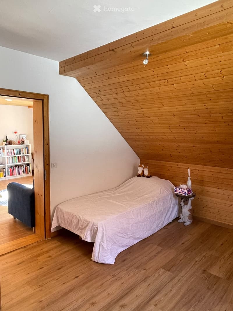 Cozy attic bedroom with wooden walls and slanted ceiling, featuring a bed, nightstand, sofa, and bookshelf in the adjacent room.