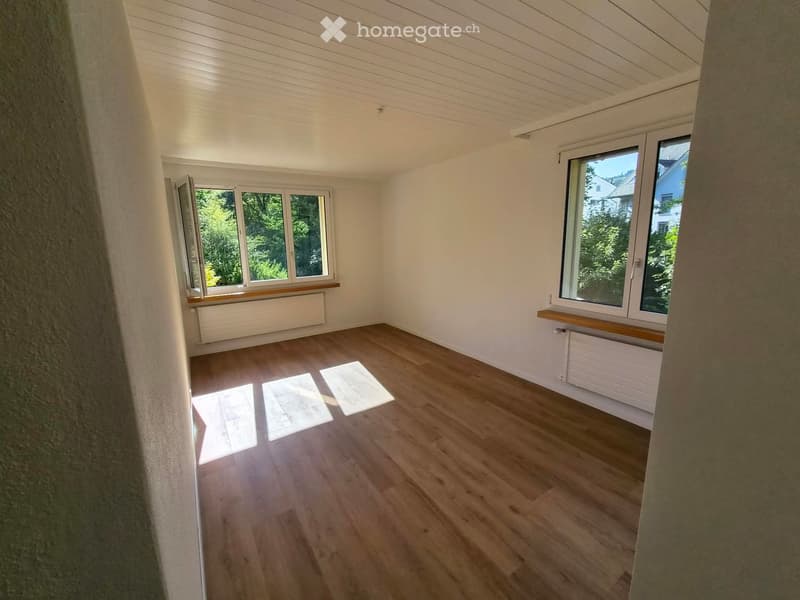 Empty room with wooden flooring, white walls, and large windows with wooden sills. Natural light streams in, casting shadows on the floor.