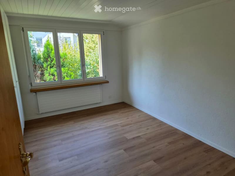 Empty room with wooden flooring, white walls, large windows, and a radiator under the windows.