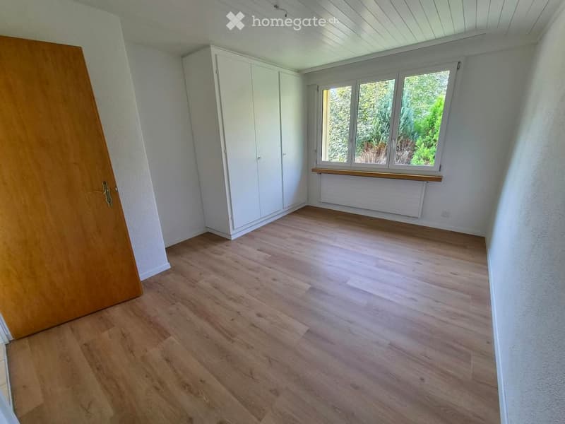 Empty room with wooden flooring, white walls, large window with wooden sill, white built-in closet, and a radiator below the window.