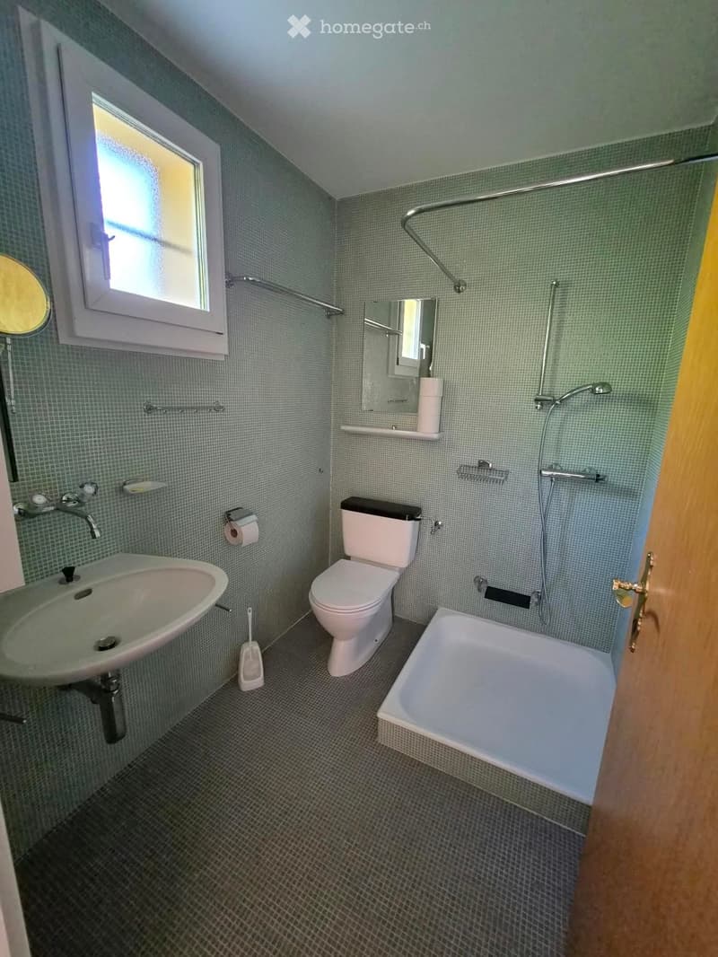 A small bathroom with a sink, toilet, bathtub, and mirror. The walls are tiled in a light green color, and there is a window providing natural light.