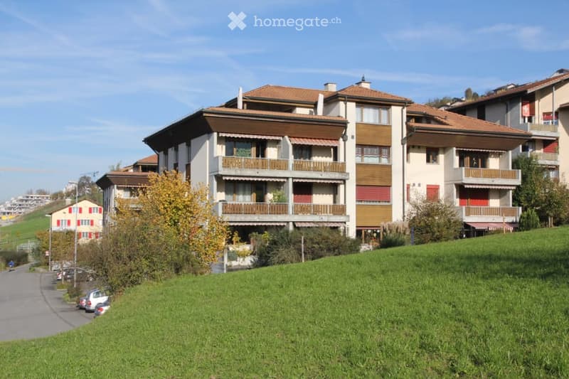 Multi-story residential building with balconies, red-tiled roof, and surrounding greenery.