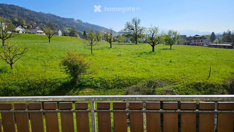 A serene view of a lush green meadow dotted with trees, with a wooden fence in the foreground and distant houses and mountains under a clear blue sky.