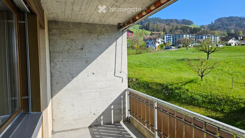 Balcony with wooden railing, overlooking a green field and distant houses, with a tree in the foreground.